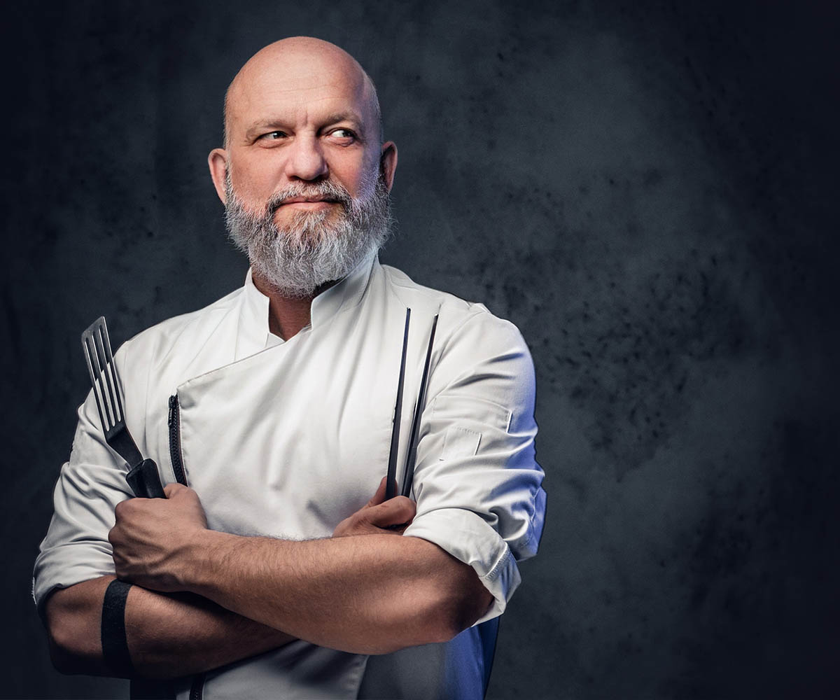 shot-of-professional-old-chef-dressed-in-uniform-holding-kitchen-spatula-and-tongs